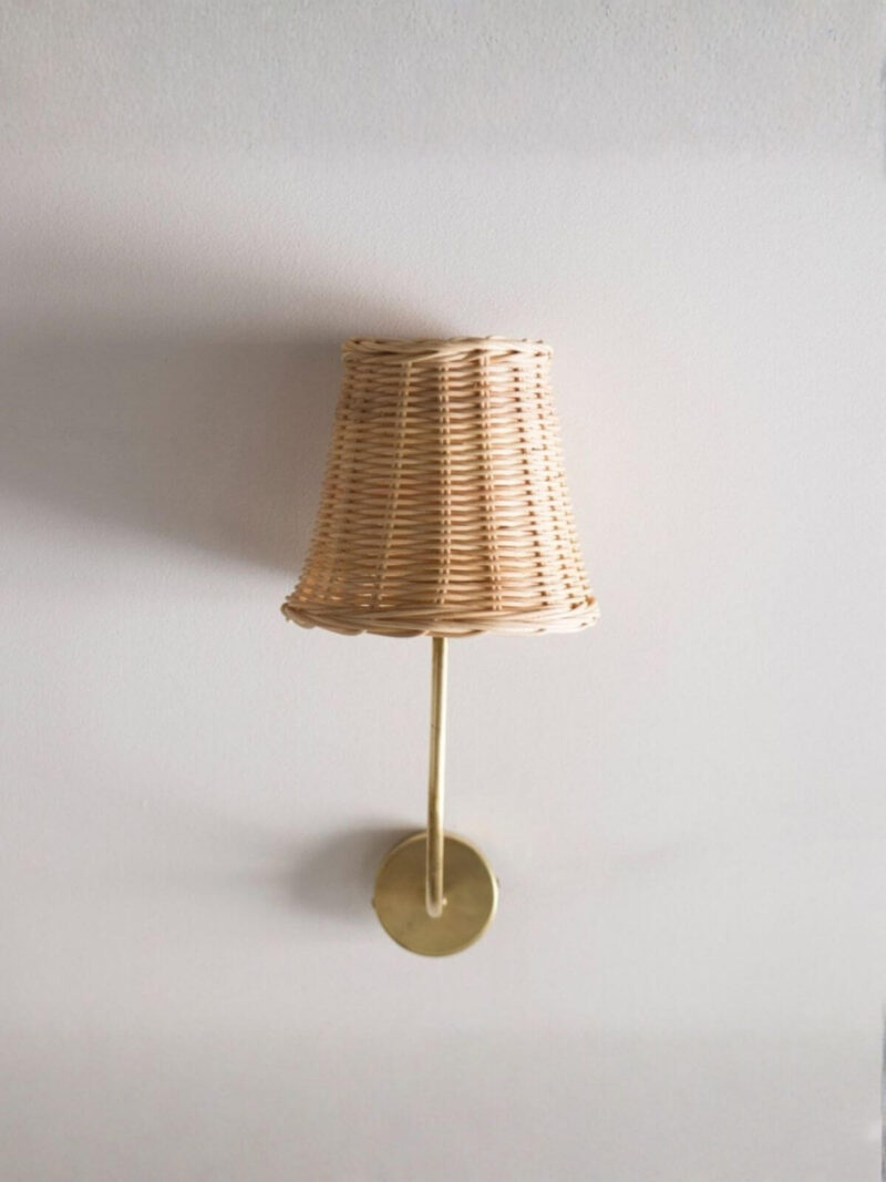 Kinkiet rattanowy ze złotym ramieniem na tle jasnej ściany.