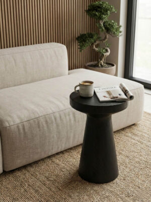 Stolik boczny klepsydra drewniany o czarnym wykończeniu ze spękanym blatem postawiony przed sofą w salonie urządzonym w stylu japandi.